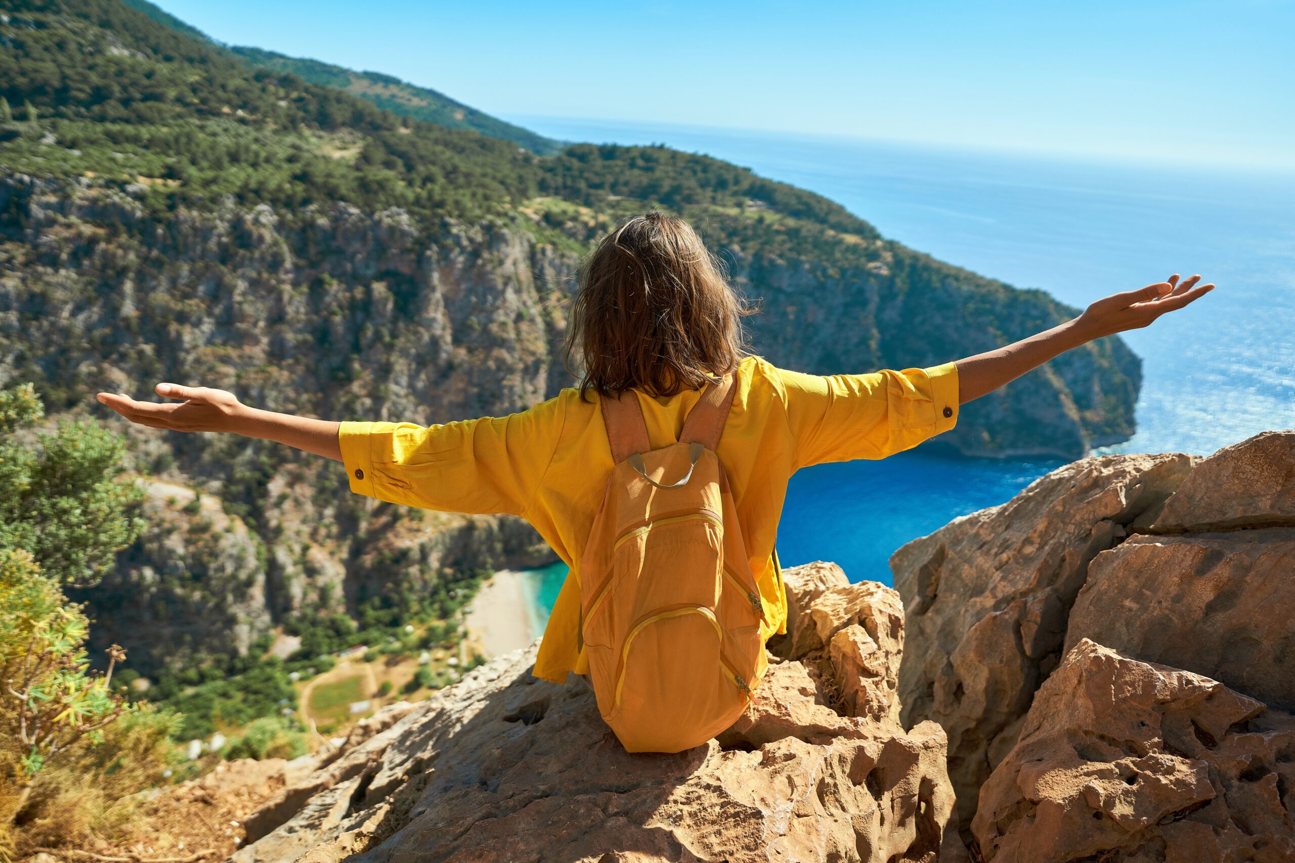 Female tourist sitting on cliff with orange backpack with open arms looking at panoramic view