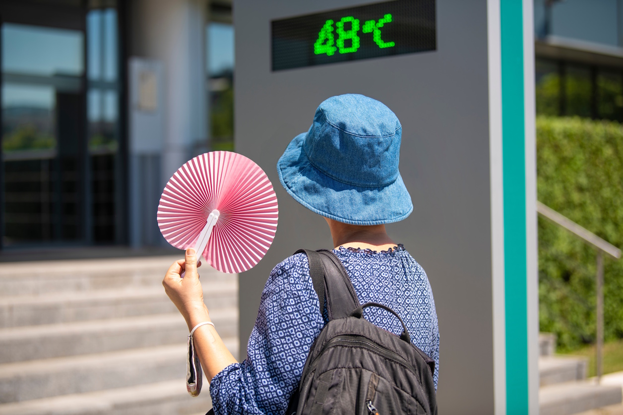 Woman wearing hat and holding paper waving fan looking at street thermometer marking 48 degrees Celsius